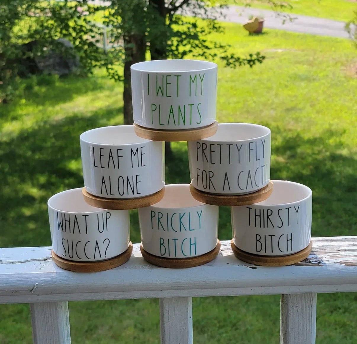 Stack of ceramic plant pots with humorous text on a wooden surface outdoors.