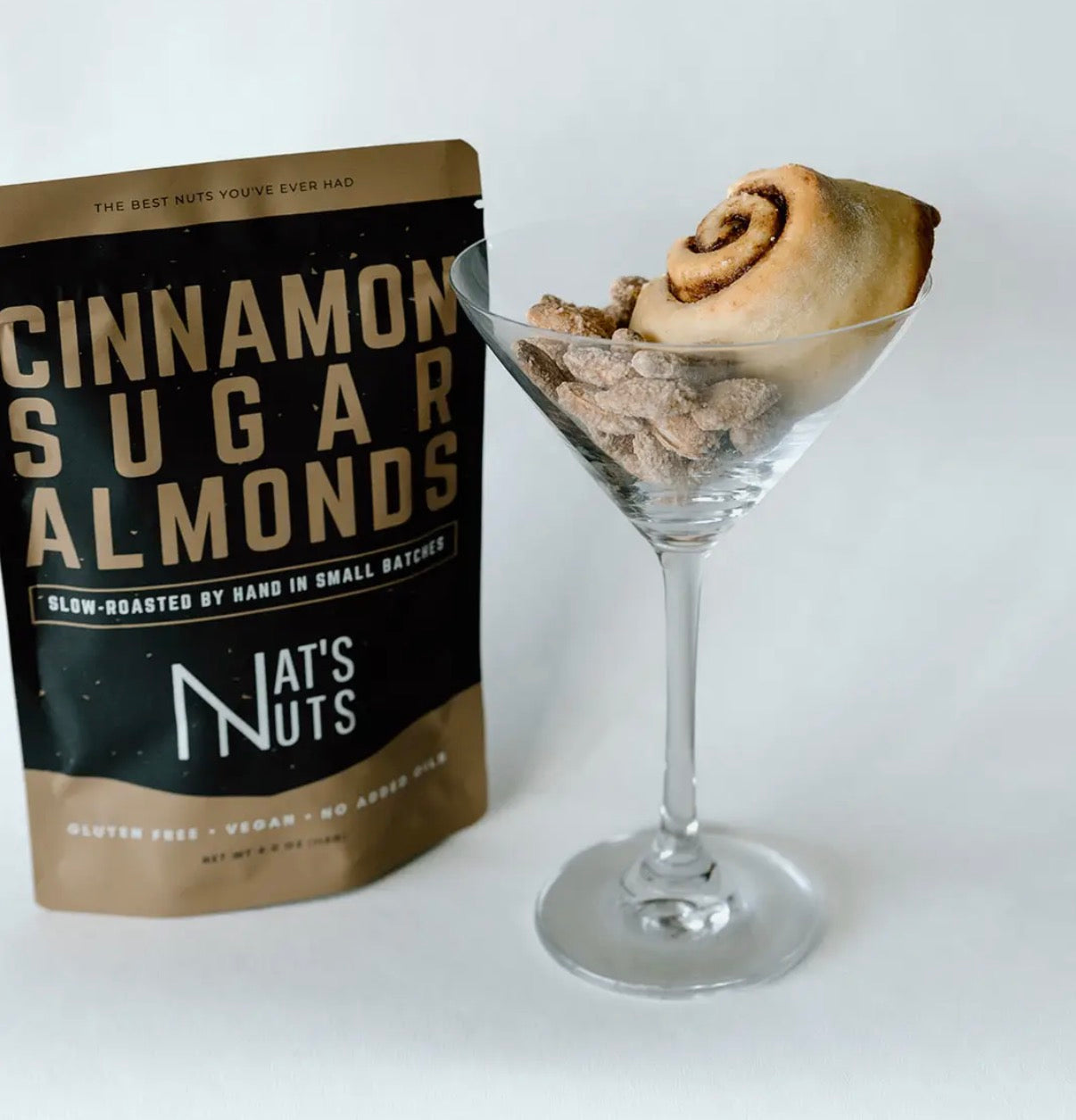 Bag of 'Cinnamon Sugar Almonds' from Nat's Nuts next to a glass with a cinnamon roll dessert on a white background