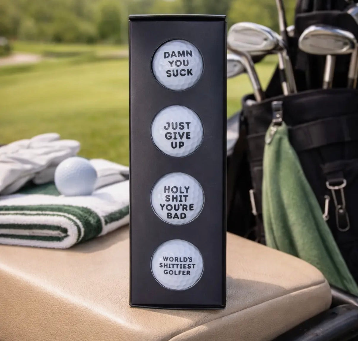 Box of golf balls with humorous text on a golf bag with golf clubs and balls in the background.
